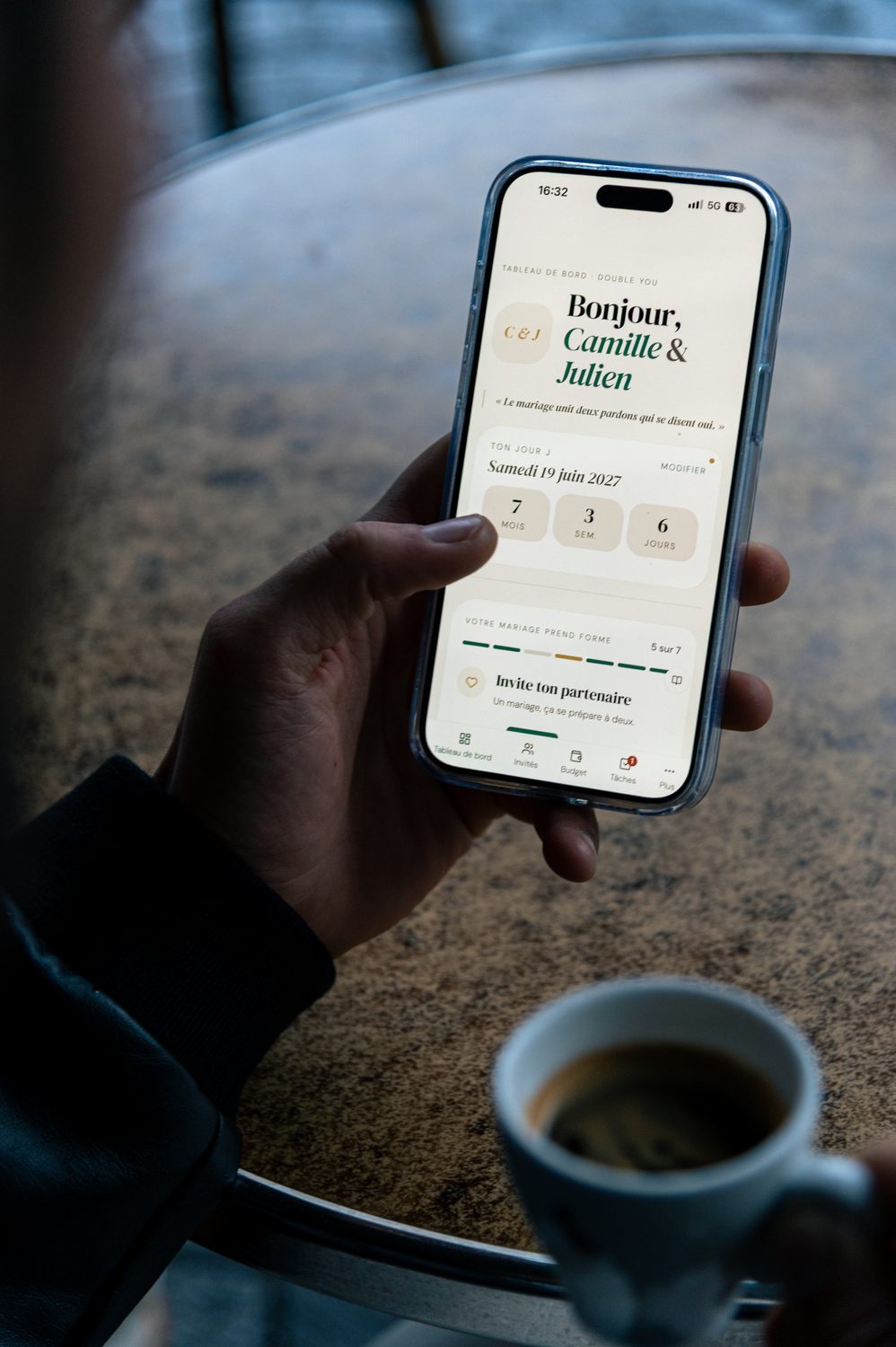 Une main tient un téléphone au-dessus d'une table de café : l'app Double You affiche « Bonjour, Camille & Julien » et le compte à rebours du mariage